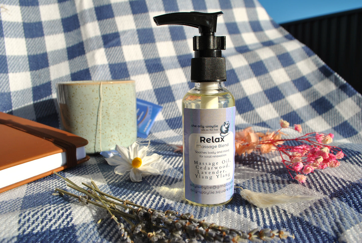Massage oil bottle labeled 'Relax' on a checkered fabric background with flowers and a notebook.