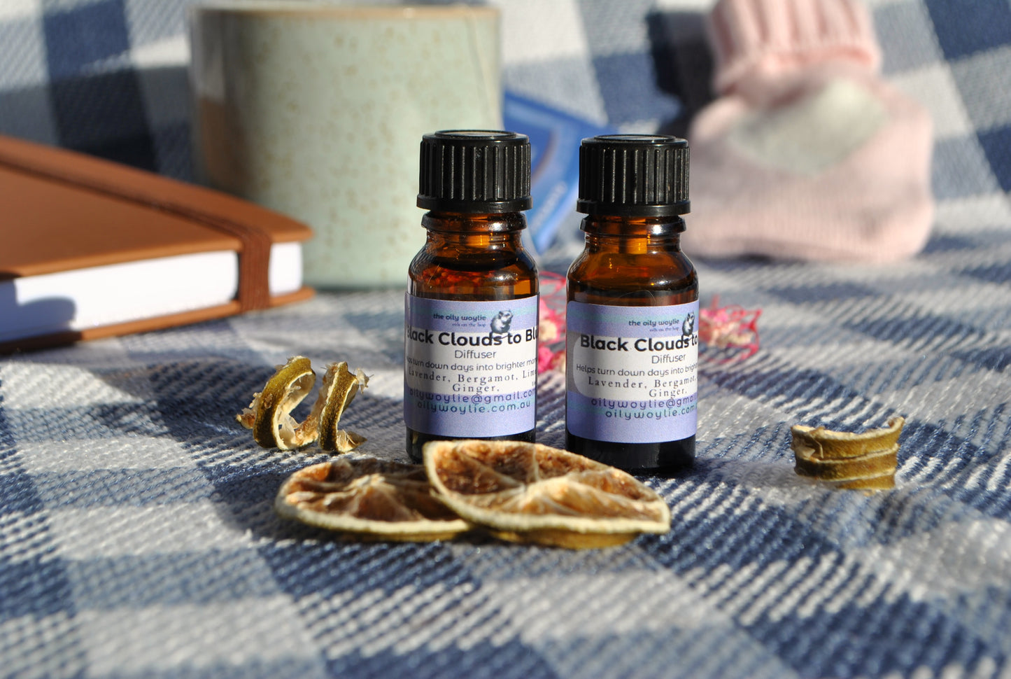 Two small bottles of 'Black Clouds to Blue' on a checkered fabric surface with dried leaves and a book.