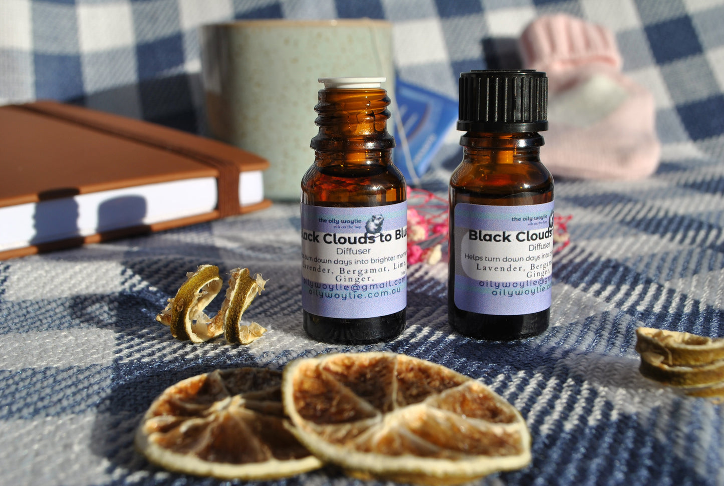 Two bottles of 'Black Clouds to Blue' diffuser on a checkered fabric background with dried oranges.