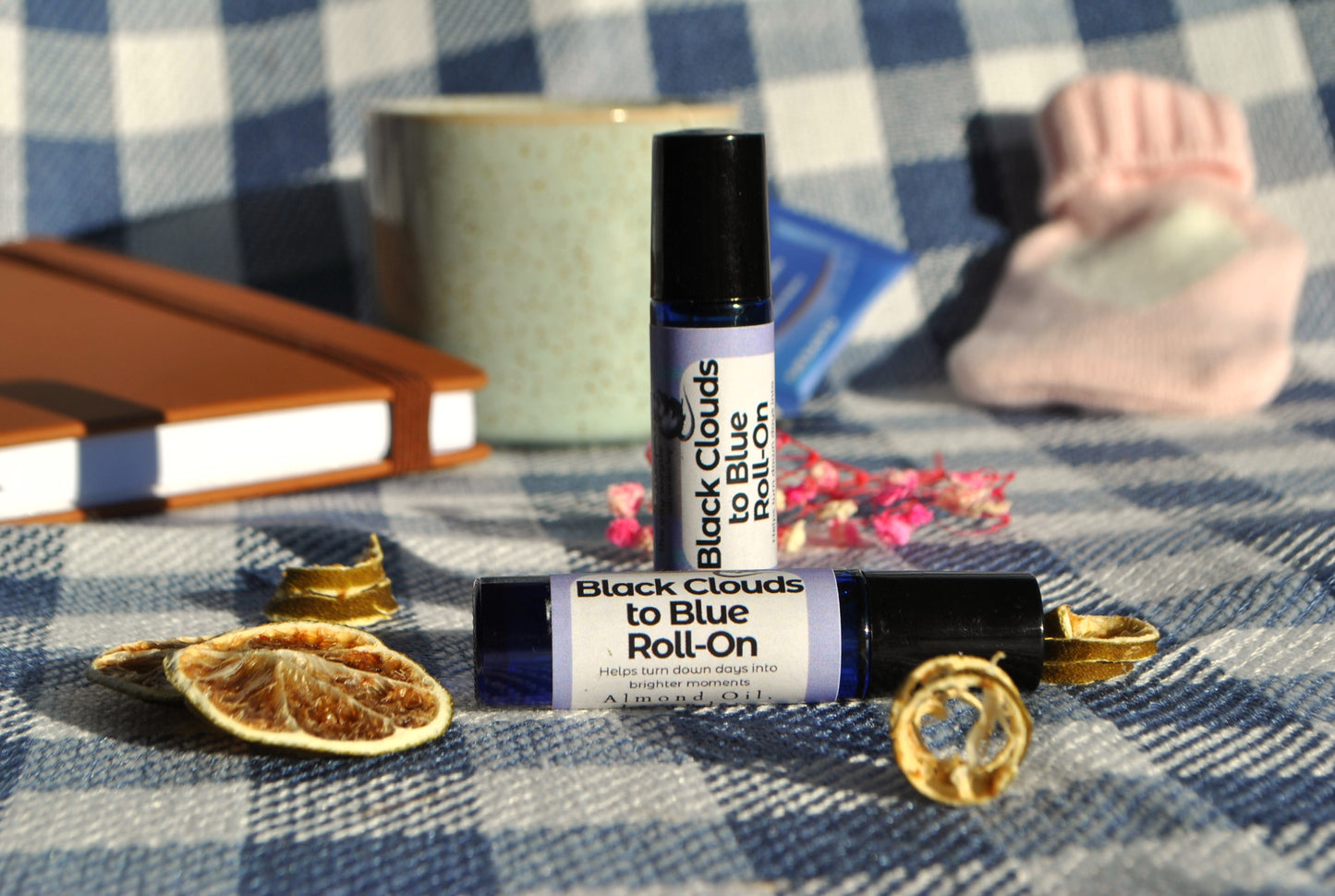 Two bottles of 'Black Clouds to Blue Roll-On' on a checkered fabric background with dried oranges and a notebook.
