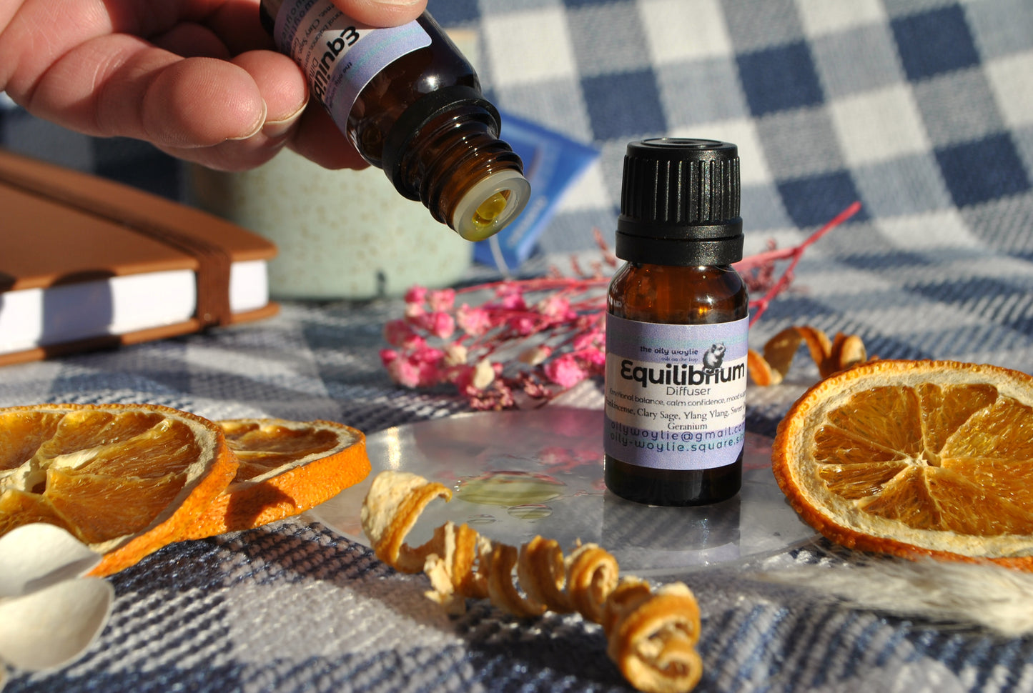 Hand holding a bottle of essential oil labeled 'Equilibrium' with dried oranges and flowers on a checkered cloth.