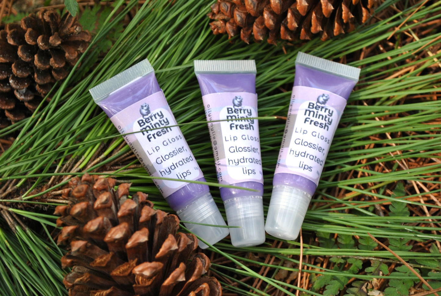 Three tubes of Berry Mint Fresh lip gloss on a background of pine cones and greenery