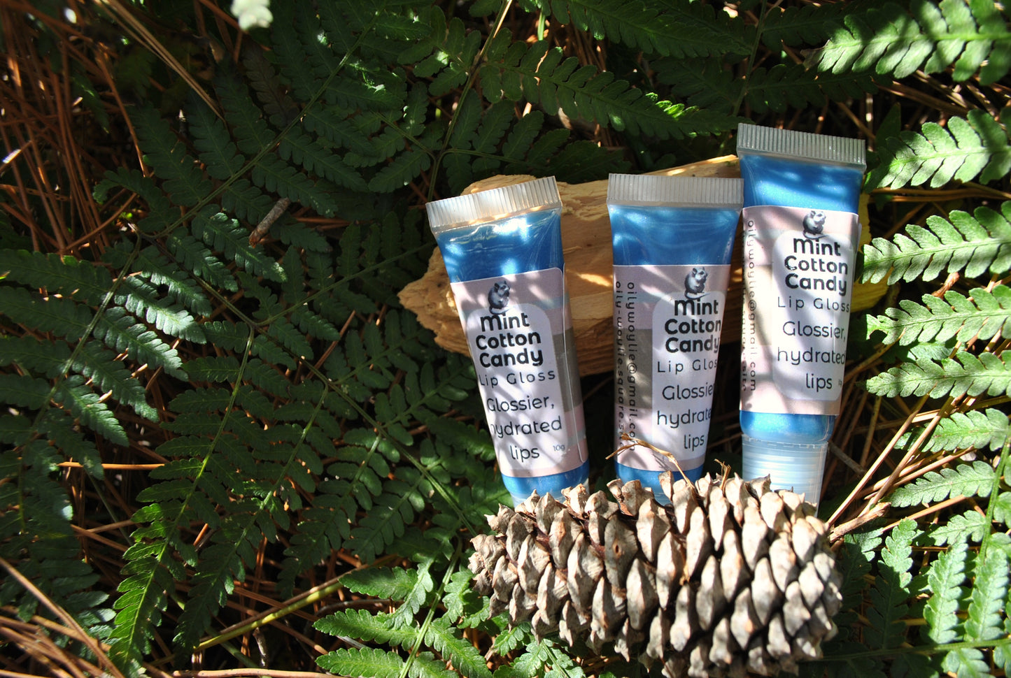 Three small bottles of mint cotton candy lip balm on a pine cone with ferns in the background