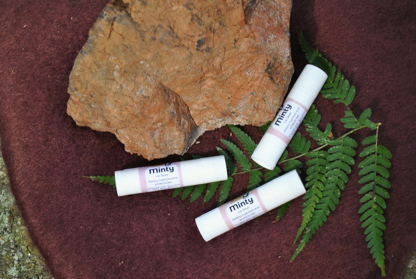 Three lip balm tubes labeled 'minty' on a rock with green leaves