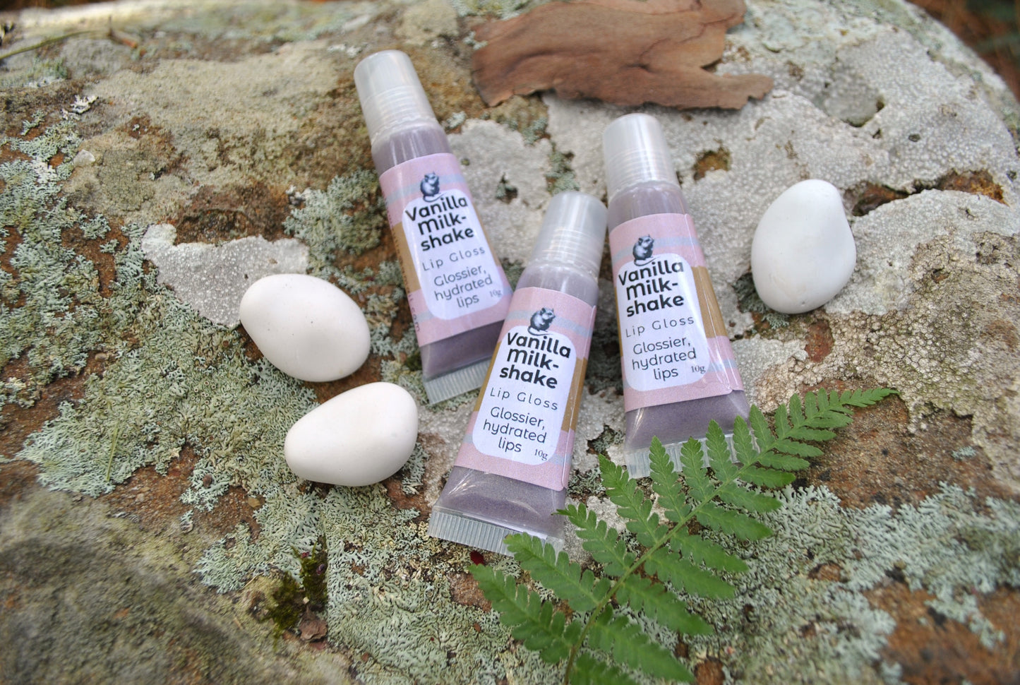 Three lip gloss bottles labeled 'Vanilla Milkshake' on a textured surface with eggs and a leaf.