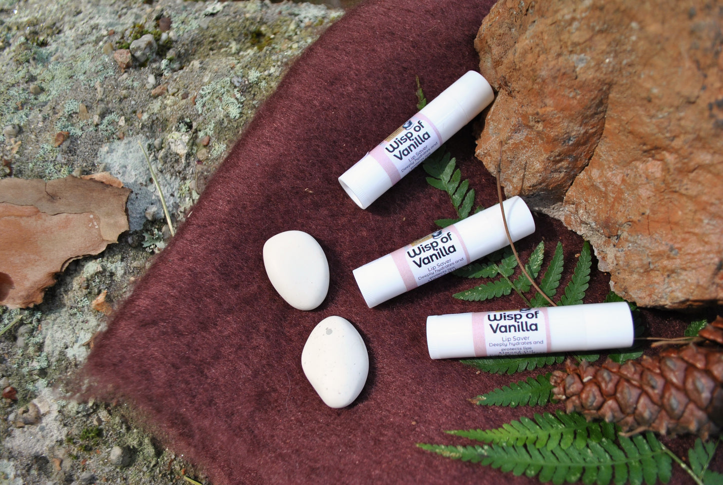 Lip balm tubes labeled 'Wisp of Vanilla' on a textured surface with leaves and rocks.
