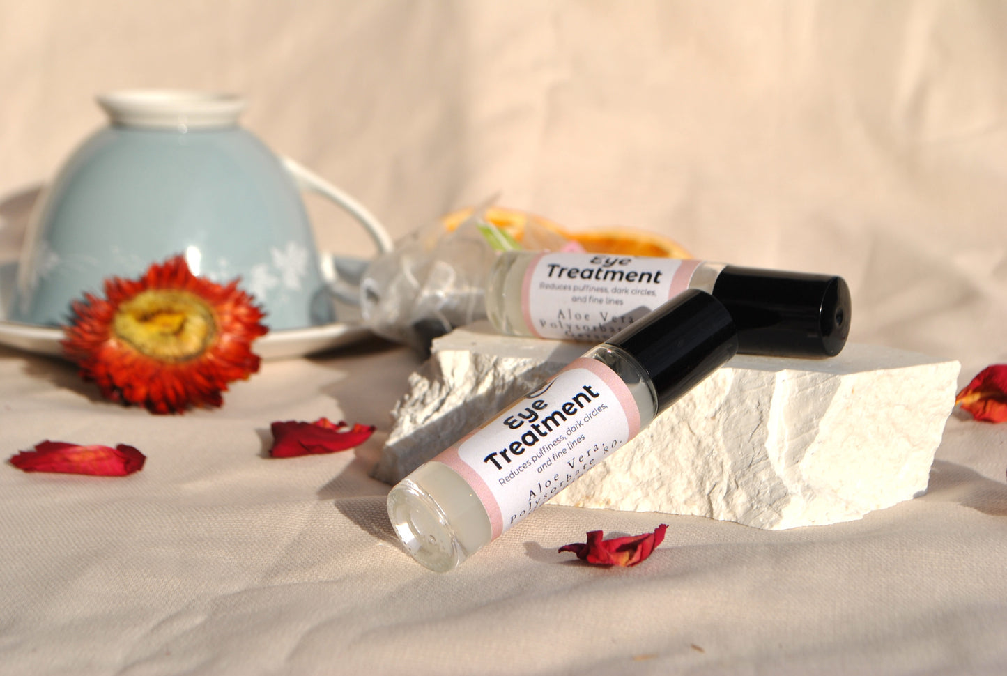 Two bottles of eye treatment on a textured surface with flowers and a teapot in the background.