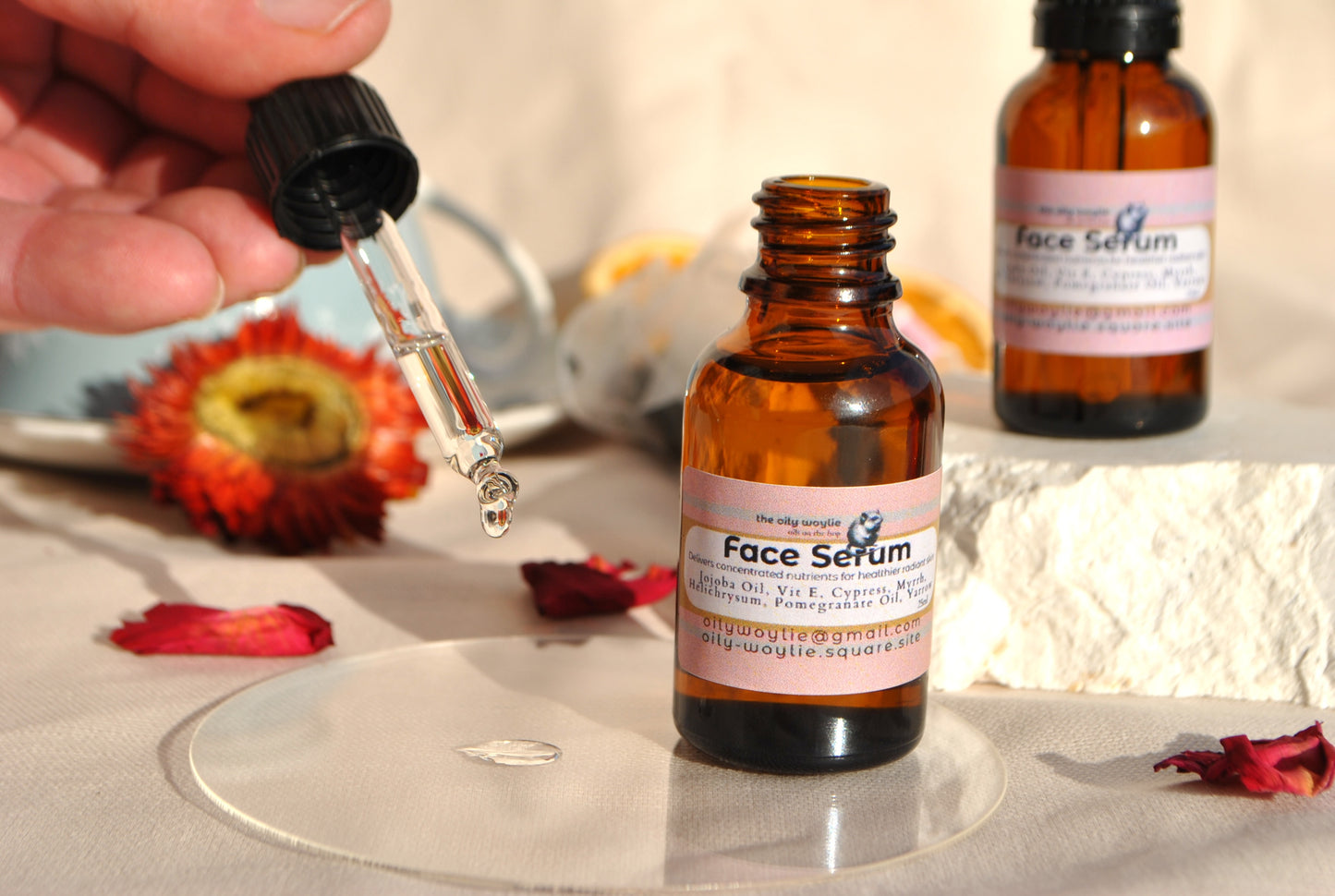 Face serum bottle with dropper on a textured surface with flowers and another bottle in the background.