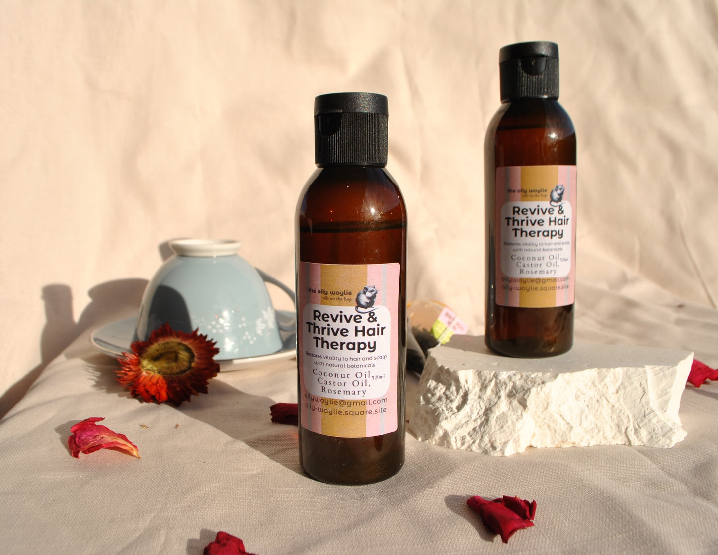 Two bottles of 'Revive & Thrive Hair Therapy' on a textured surface with flowers and a teapot in the background.