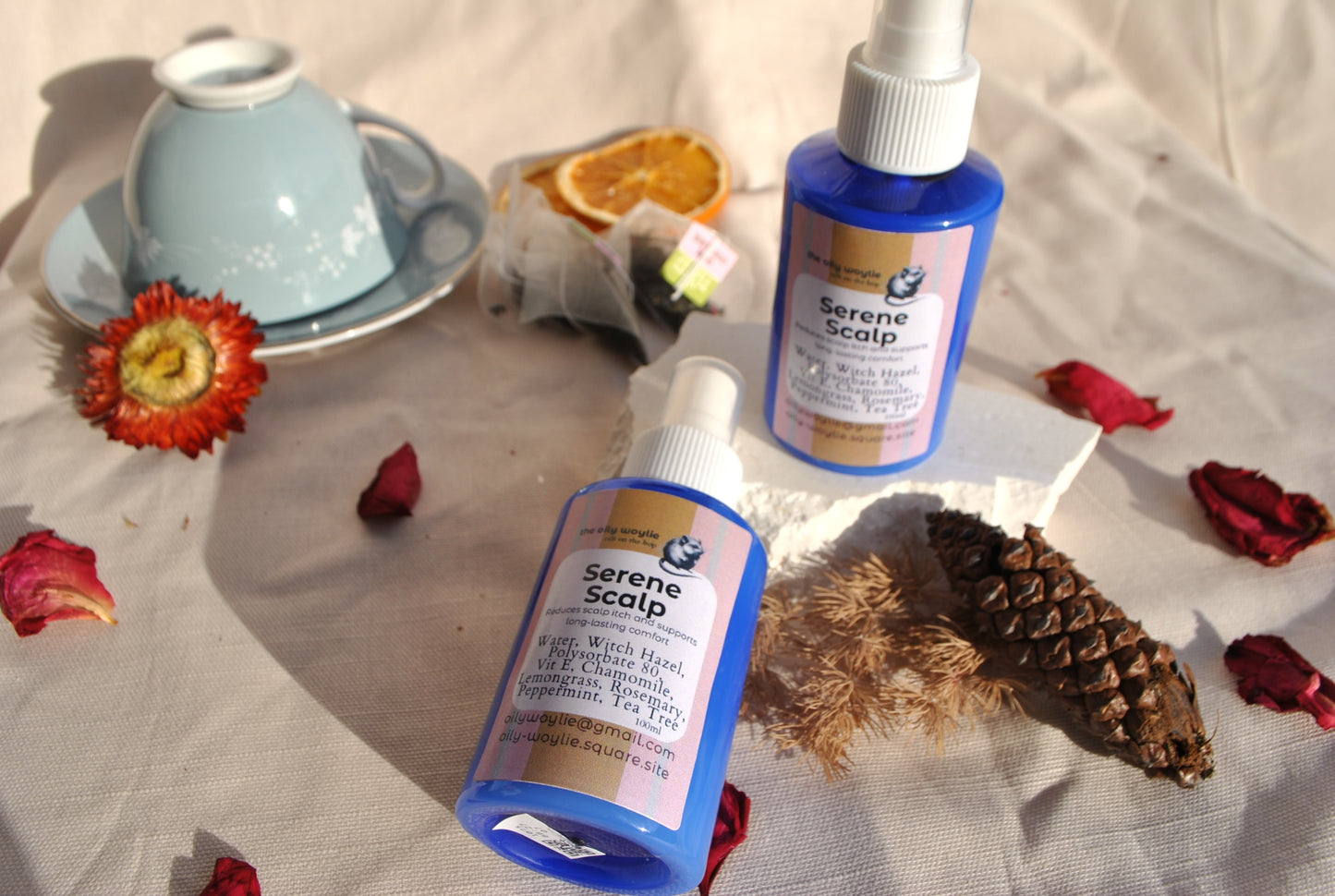 Two bottles of 'Serene Scalp' product on a textured surface with decorative elements like a teacup, flowers, and pinecones.