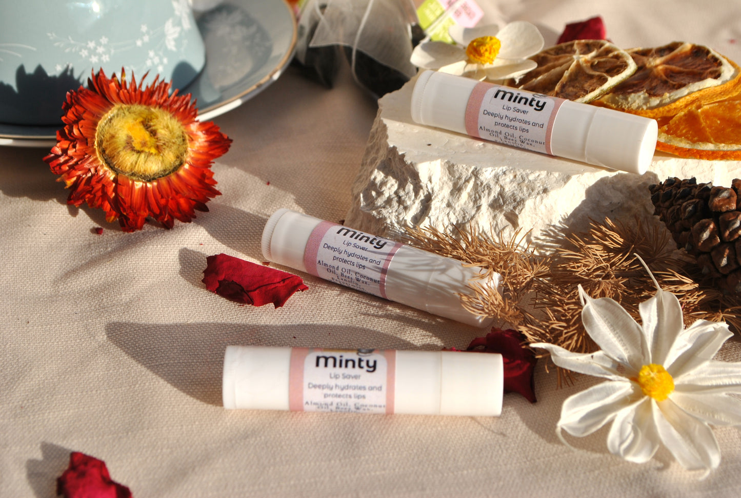 Minty lip balm tubes on a textured surface with flowers and a bowl in the background