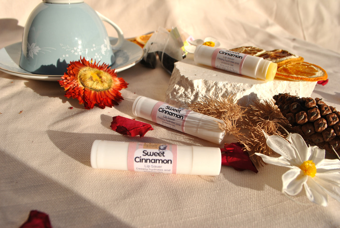 Lip balm sticks labeled 'Sweet Cinnamon' on a table with decorative items including a teacup and flowers.