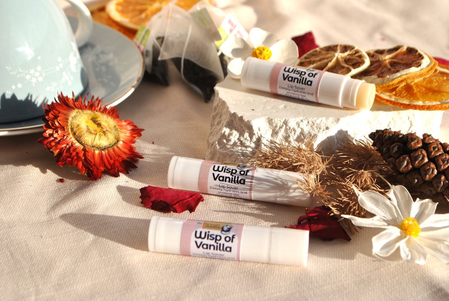 Three 'Wisp of Vanilla' lip balms on a textured surface with flowers and a teacup in the background.