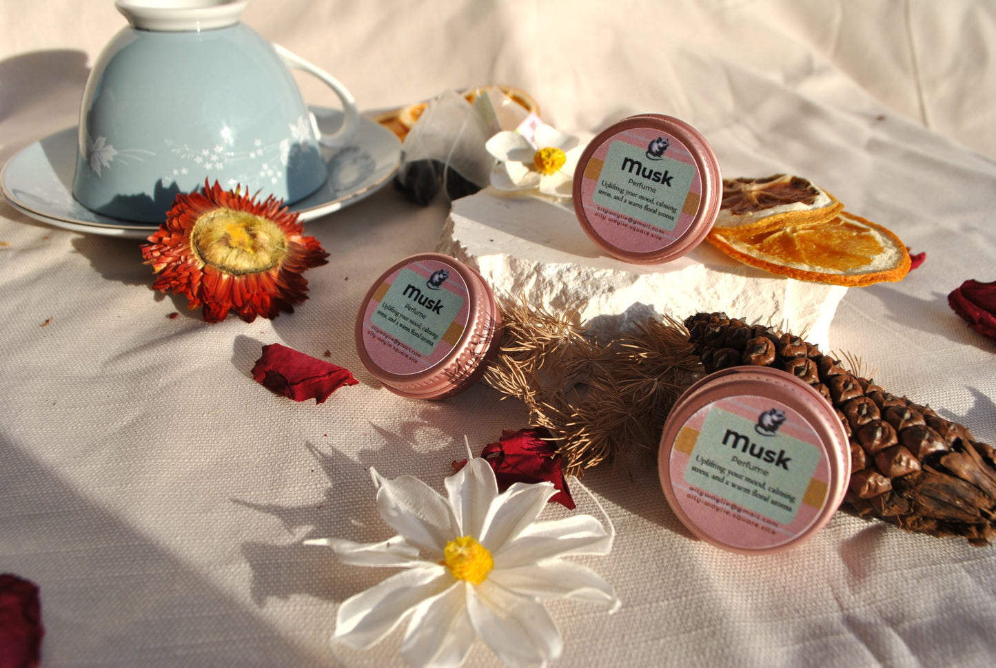 Three pink containers labeled 'Musk' on a table with flowers and pine cones.