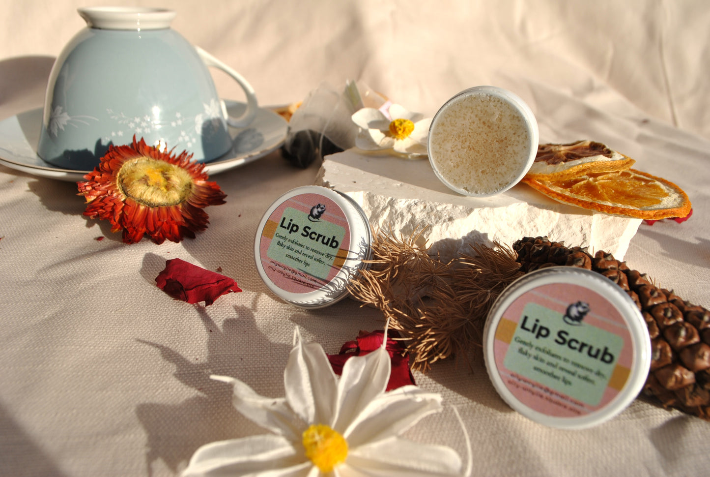 Lip scrub containers on a table with flowers and a teacup in the background