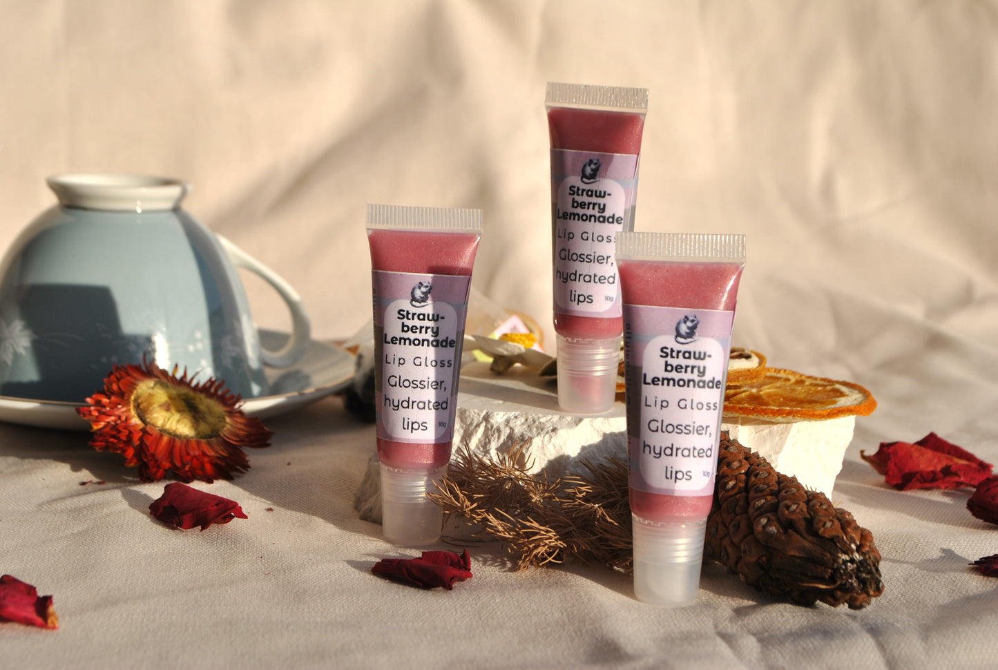 Three tubes of strawberry lemonade lip gloss on a textured surface with dried flowers and a teacup in the background.