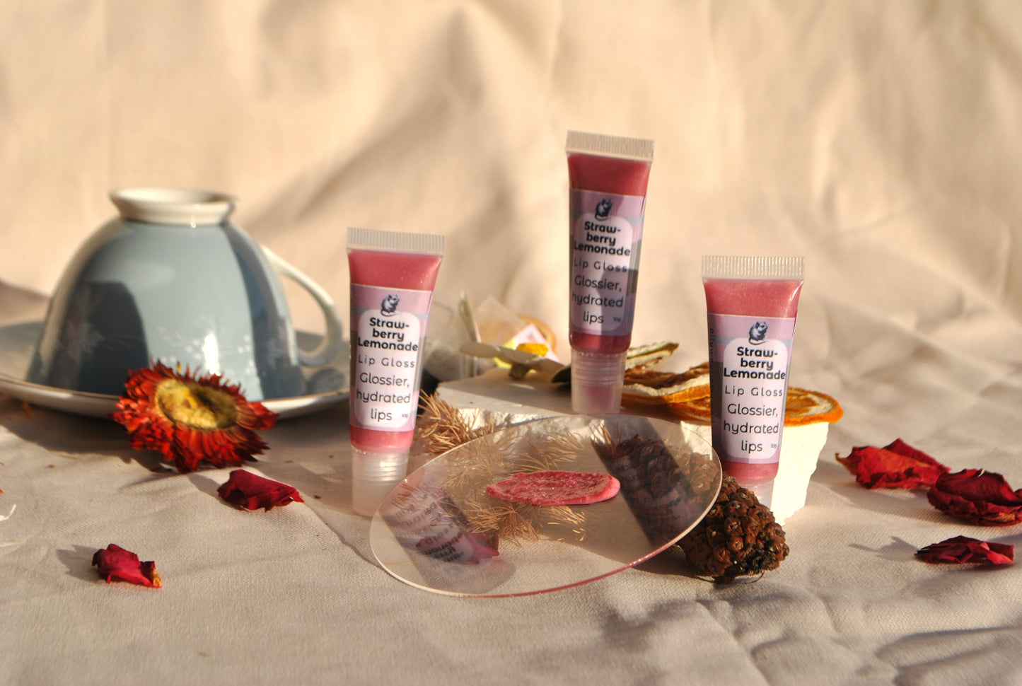 Three tubes of strawberry lemonade lip gloss on a decorative surface with flowers and a candle.