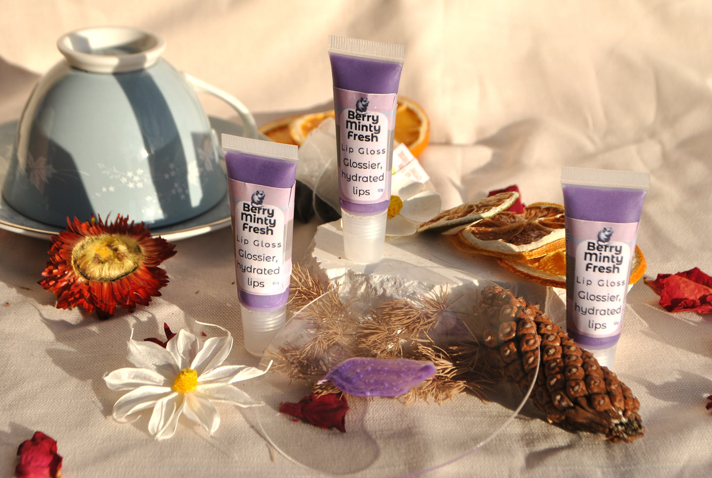 Three tubes of Berry Minty Fresh lip gloss on a decorative surface with flowers and a silver bowl.