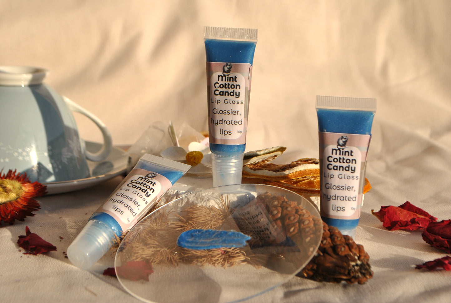 Three tubes of Mint Cotton Candy lip balm on a textured surface with flowers and a teacup in the background.