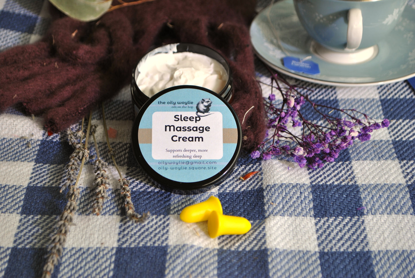 Jar of Sleep Massage Cream on a checkered tablecloth with a cup and flowers.