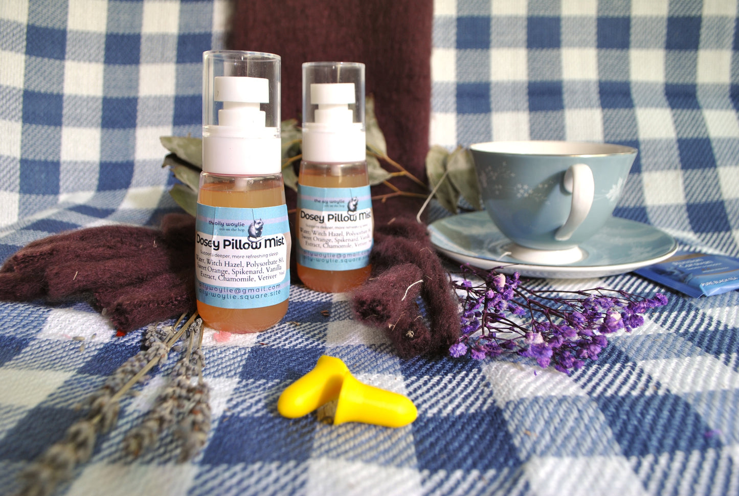 Two bottles of 'Dosey pillow mist' on a checkered tablecloth with a cup, saucer, and lavender flowers.