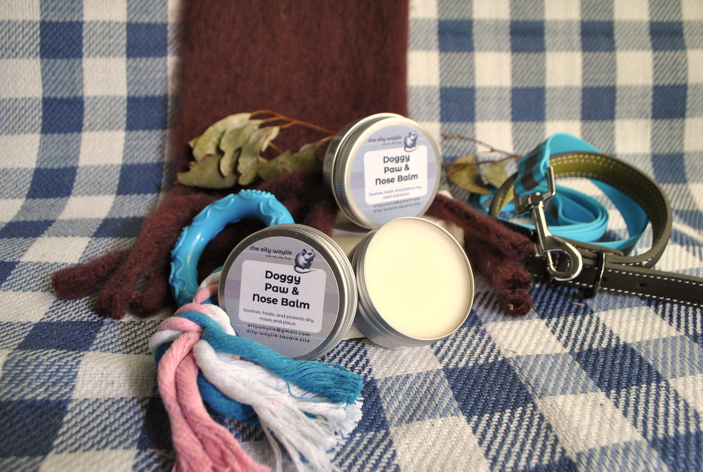 Doggy Paw & Nose Balm tins with a dog leash and bandana on a checkered fabric background