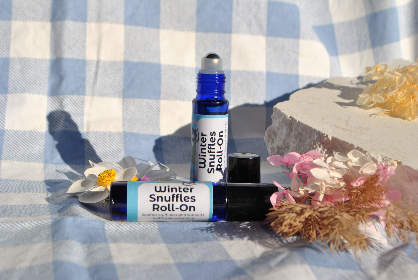 Two roller bottles labeled 'Winter Snuffles Roll On' on a checkered fabric background with flowers.