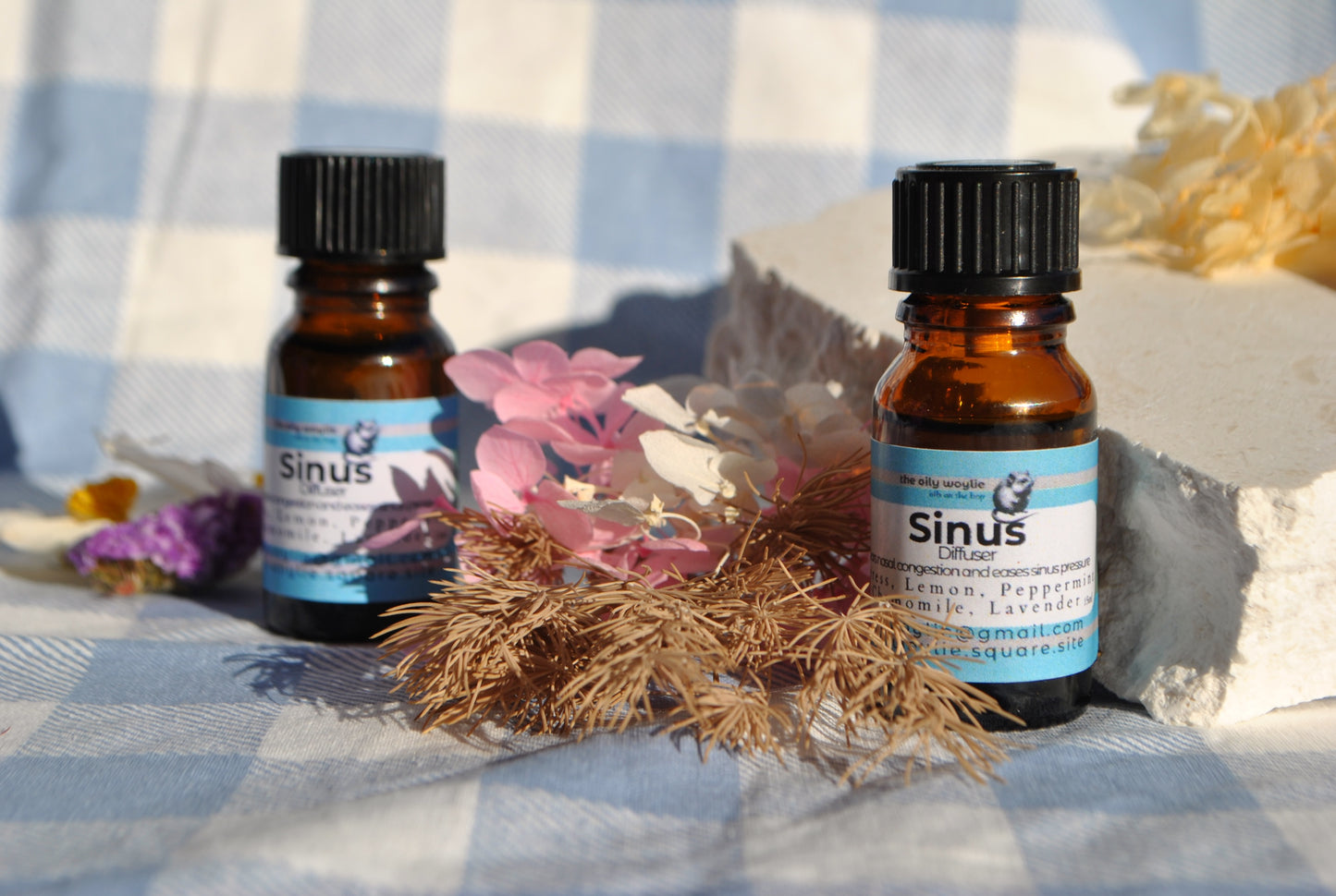 Two bottles of 'Sinus' essential oil on a checkered fabric background with flowers and herbs.