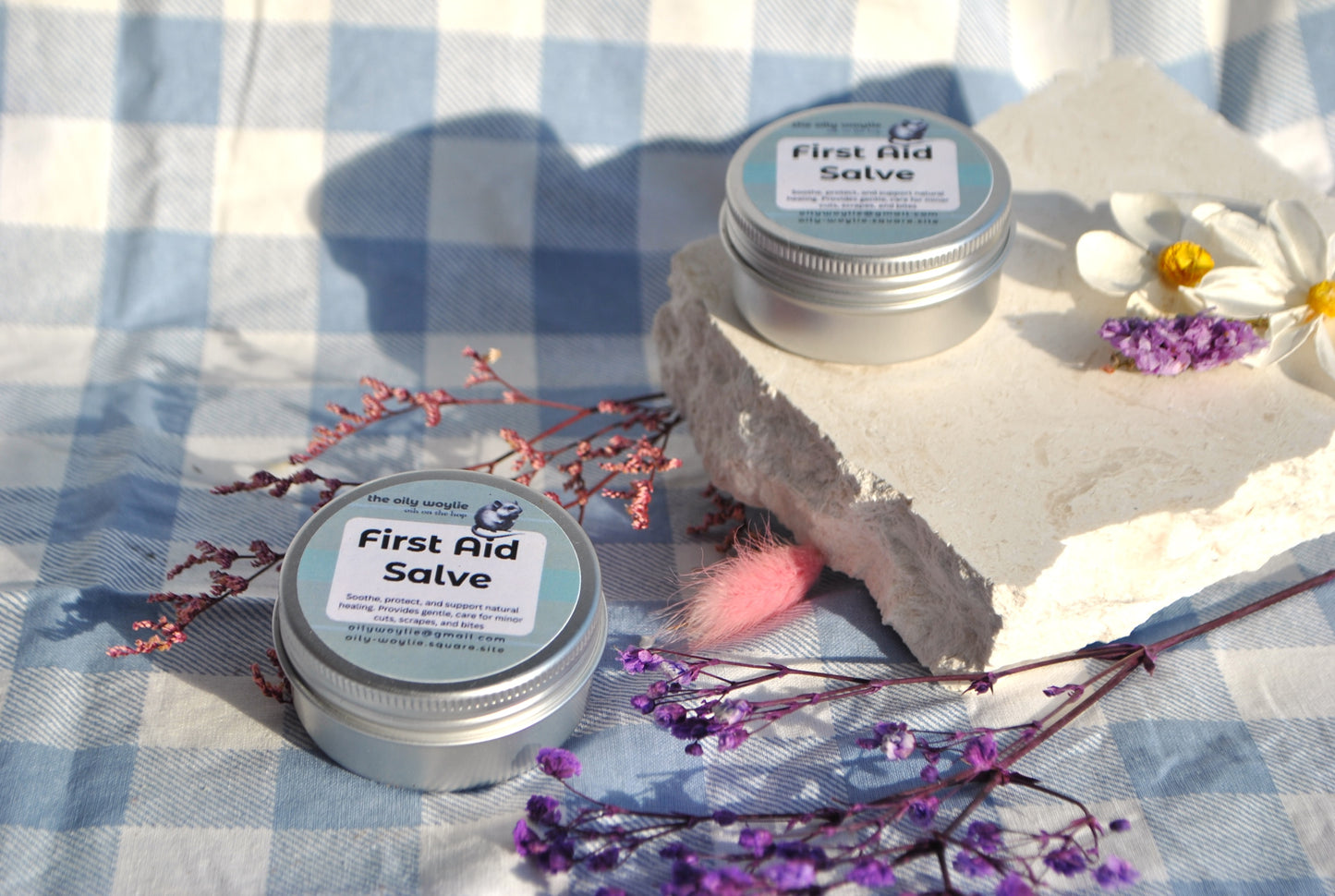 Two tins of 'First Aid Salve' on a stone surface with flowers and a checkered cloth background.