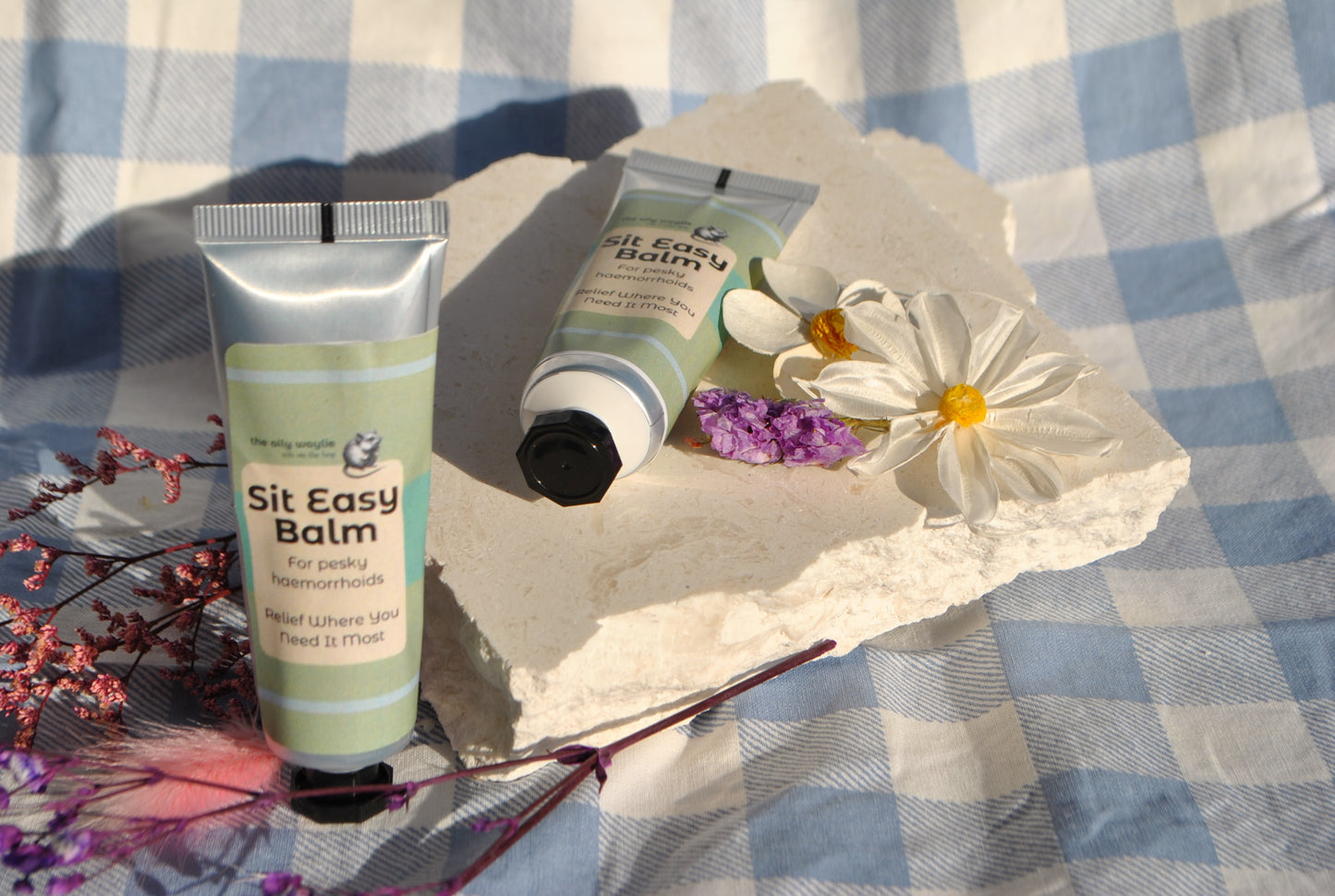 Two bottles of 'Sit Easy Balm' on a textured surface with flowers and leaves.