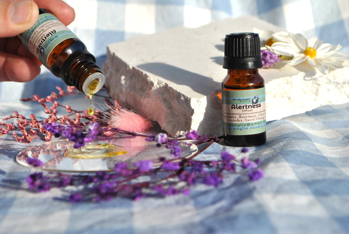 Two bottles of essential oil labeled 'Alertness' on a reflective surface with flowers and a checkered cloth background.