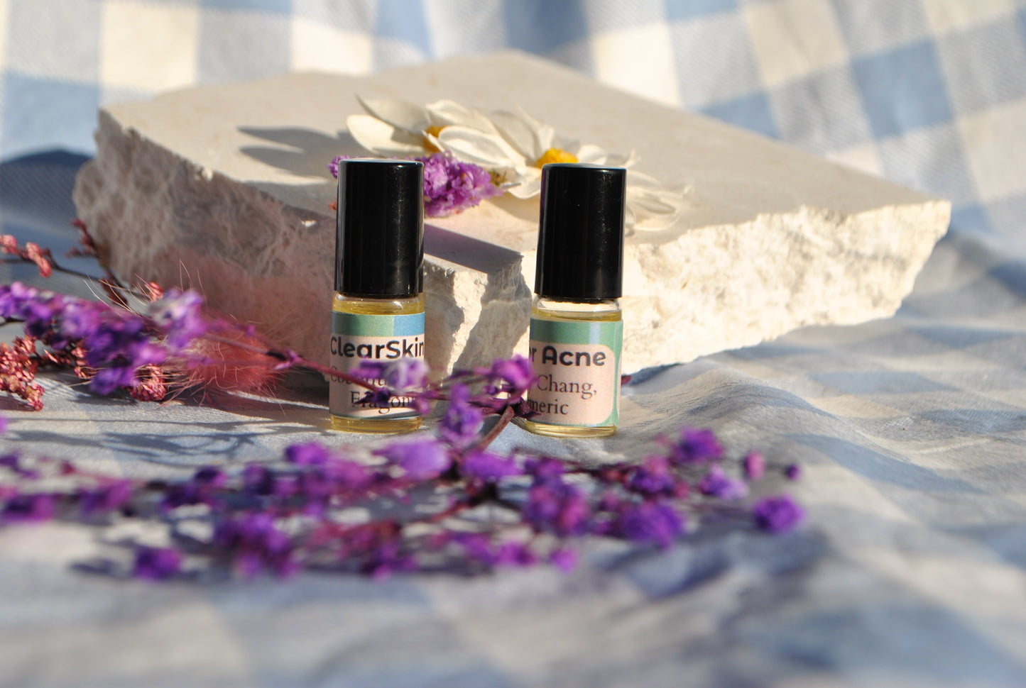 Two small bottles labeled 'Clear Skin' and 'Acne' on a stone surface with purple flowers.