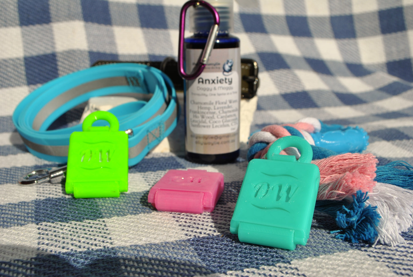 Dog training equipment including a leash, clicker, and anxiety medication bottle on a checkered blanket.