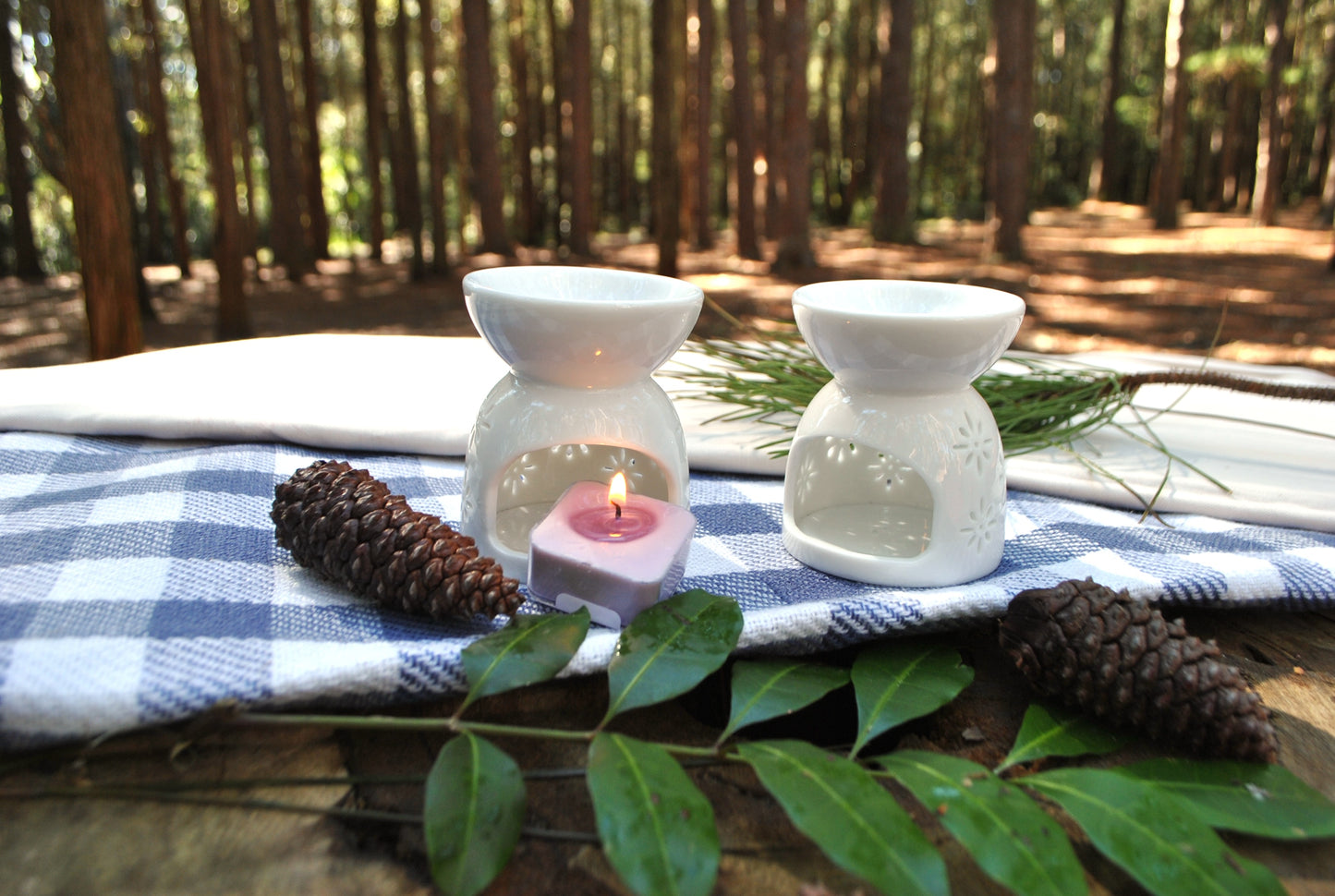 Two white ceramic oil burners with a lit pink candle on a checkered cloth outdoors.