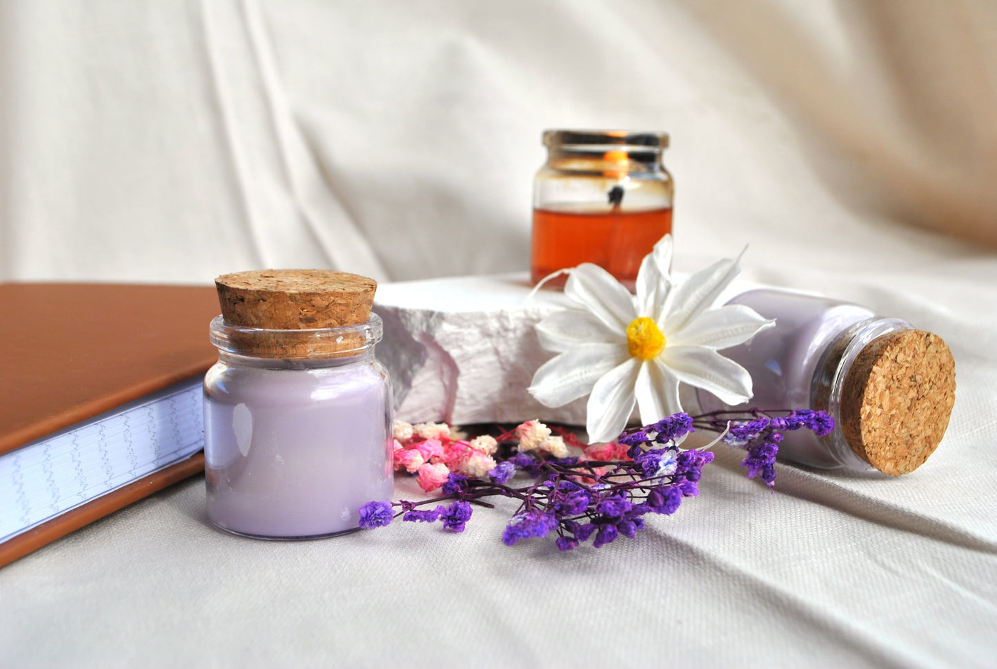 Two small jars with cork lids, a book, and flowers on a light fabric background