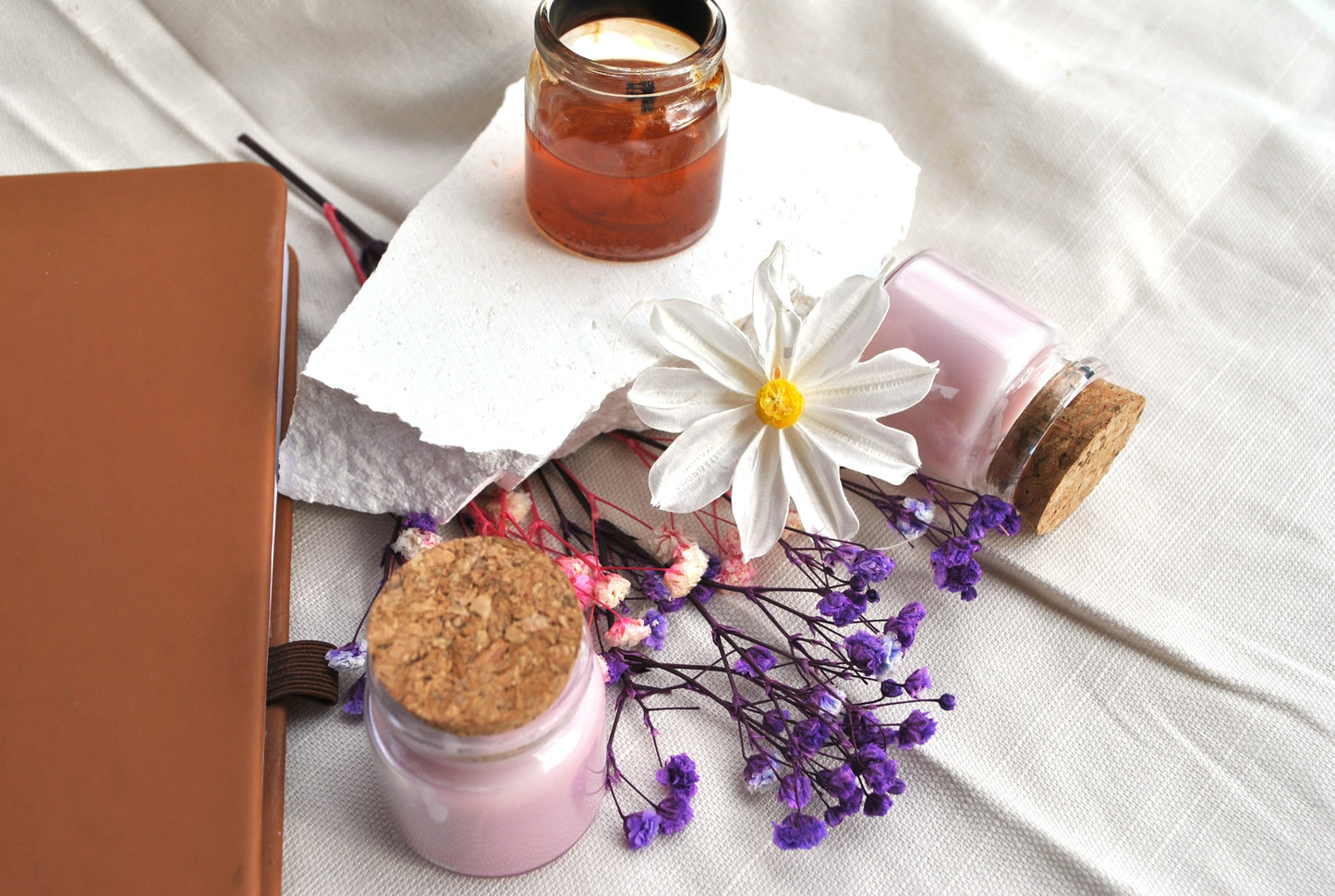 Three small jars with cork lids on a white surface with flowers and a notebook.