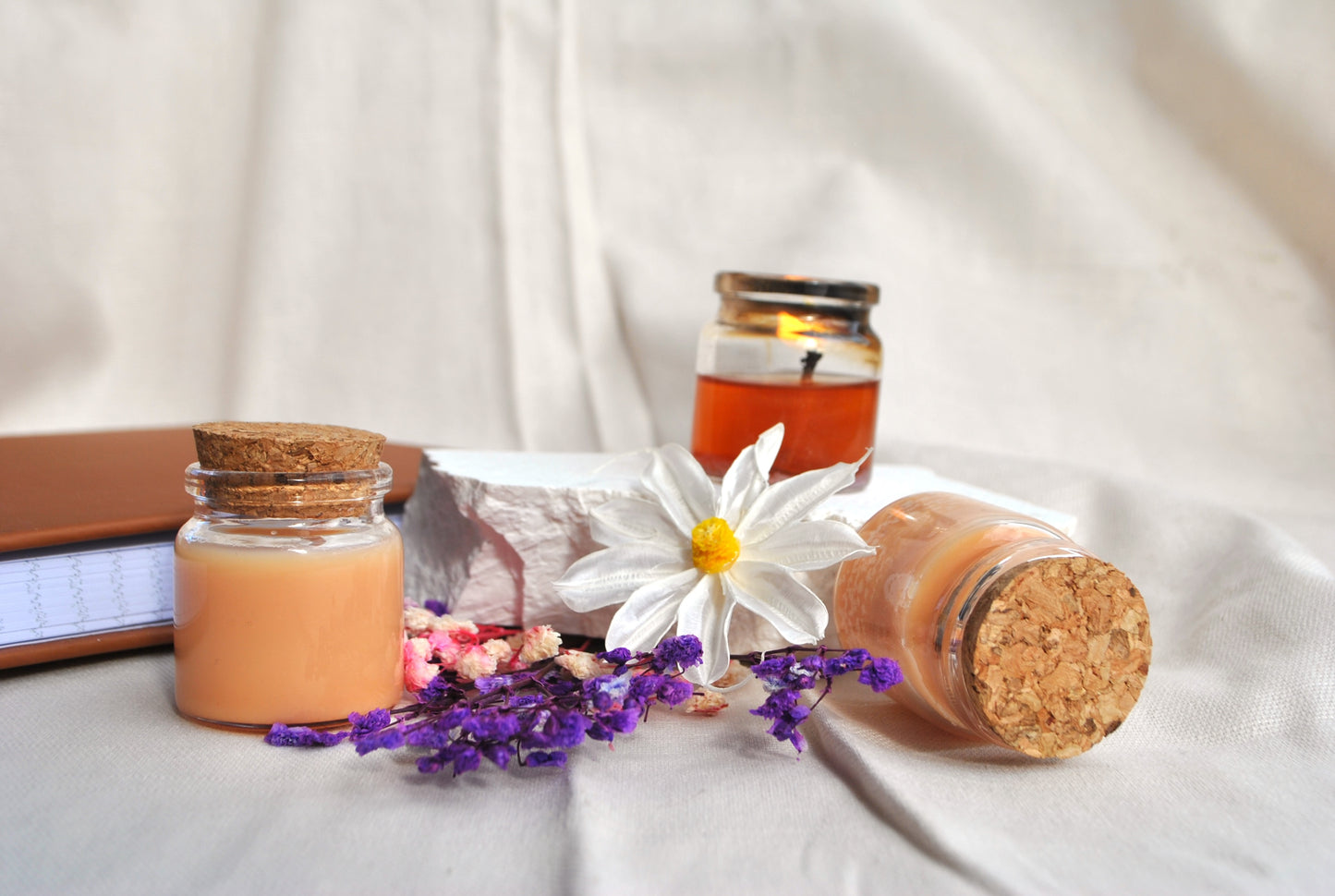 Three small jars with cork lids, a book, and a white flower on a light fabric background
