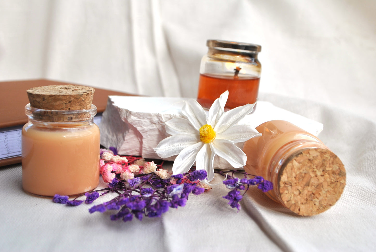 Three small jars with cork lids, a book, and flowers on a white surface