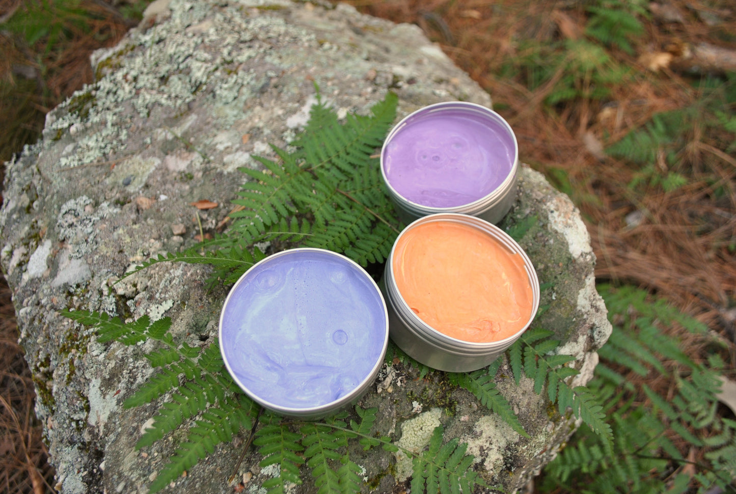 Three tins of bath salts, two purple and one orange, on a rock with greenery.