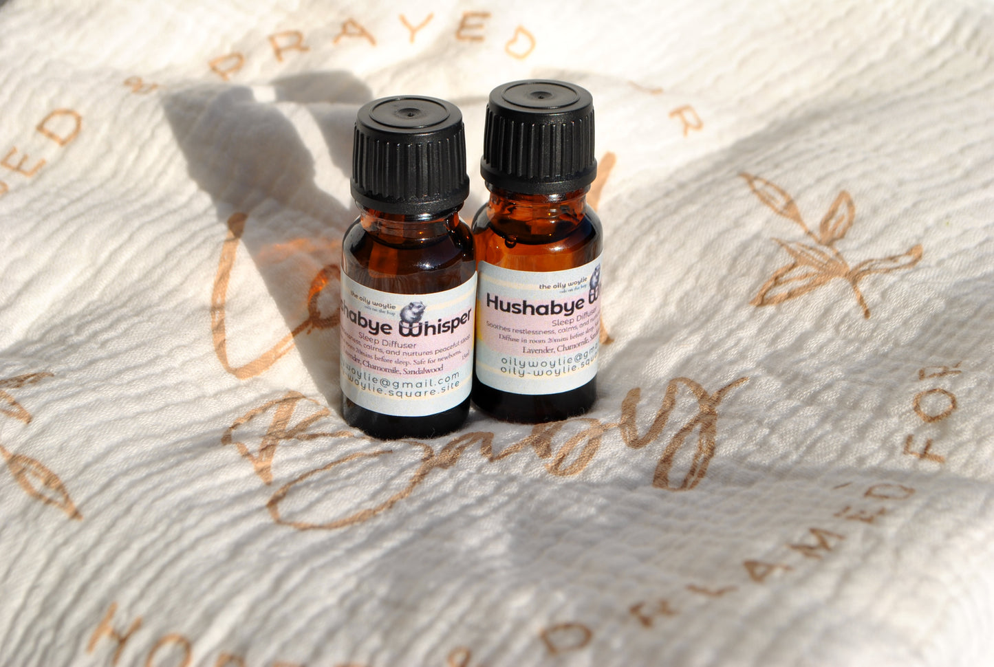 Two small bottles of 'Hushabye' and 'Baby Whisper' on a textured fabric background.