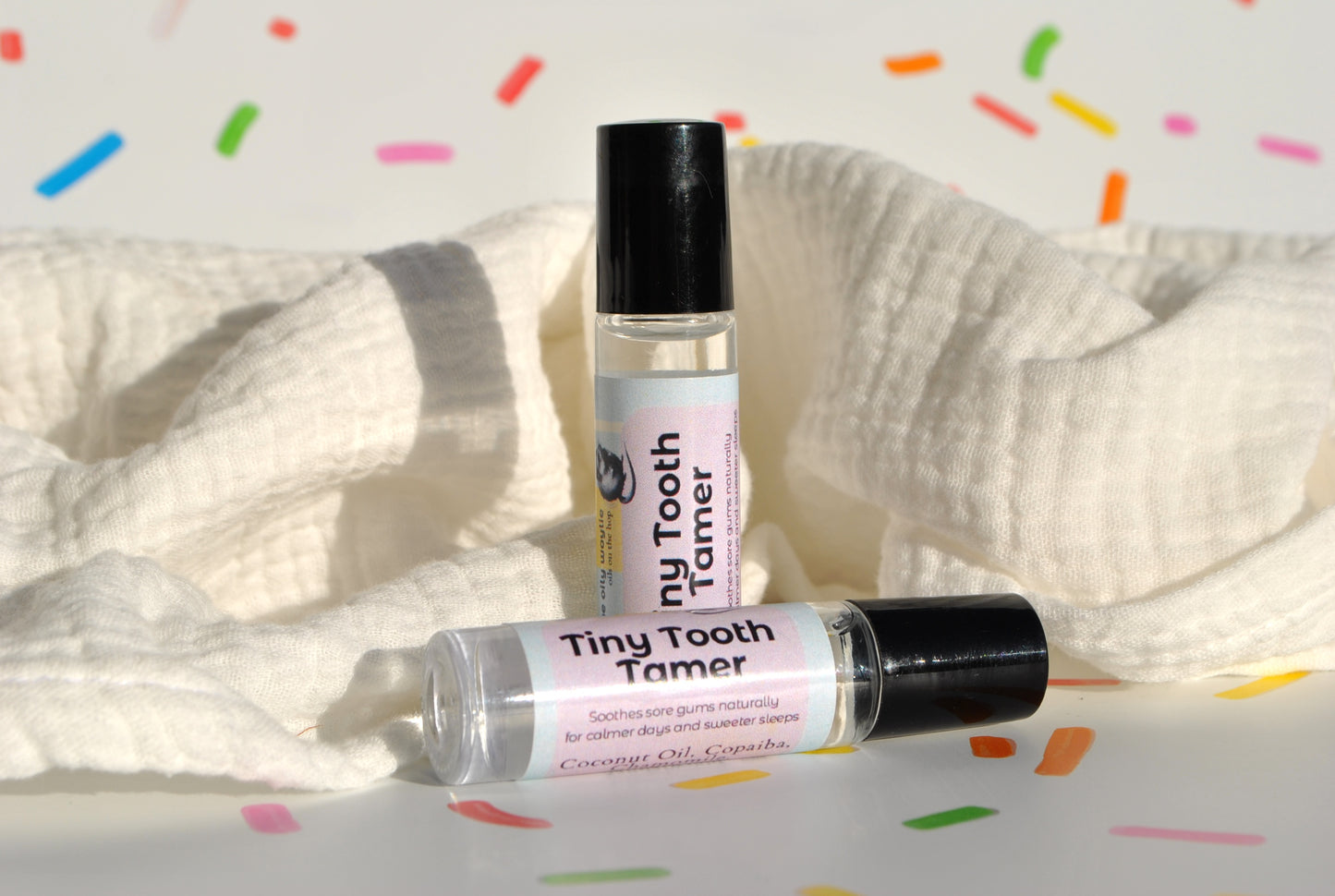 Two small roller bottles labeled 'Tiny Tooth Tamer' on a white background with colorful confetti.