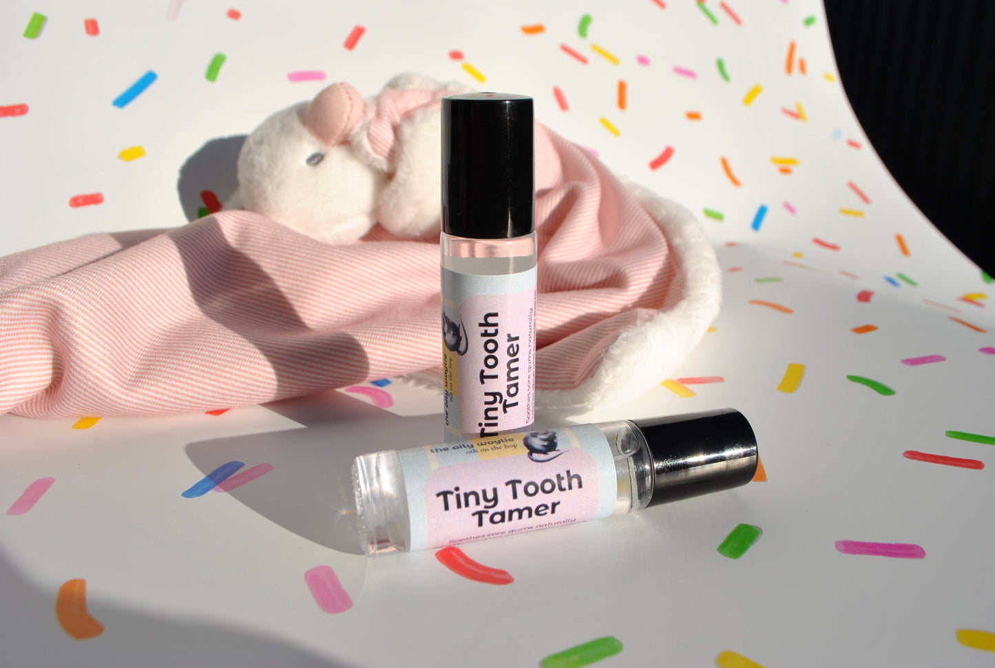 Two bottles of 'Tiny Tooth Tamer' on a surface with colorful sprinkles in the background.