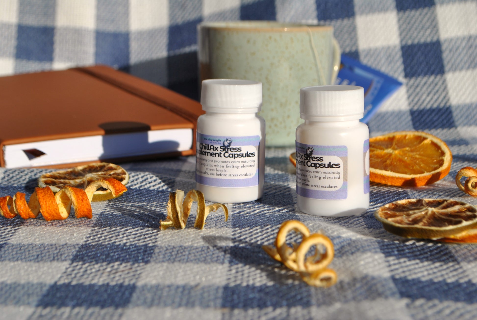 Two bottles of 'Chillax Stress' supplement capsules on a checkered tablecloth with dried oranges and a notebook.