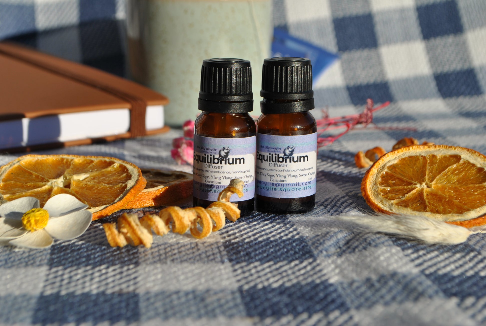 Two bottles of Equilibrium essential oils on a checkered tablecloth with dried oranges and flowers.
