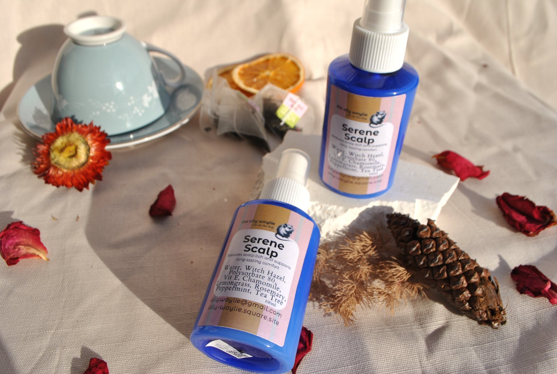 Two bottles of 'Serene Scalp' product on a textured surface with decorative elements like a teacup, flowers, and pinecones.