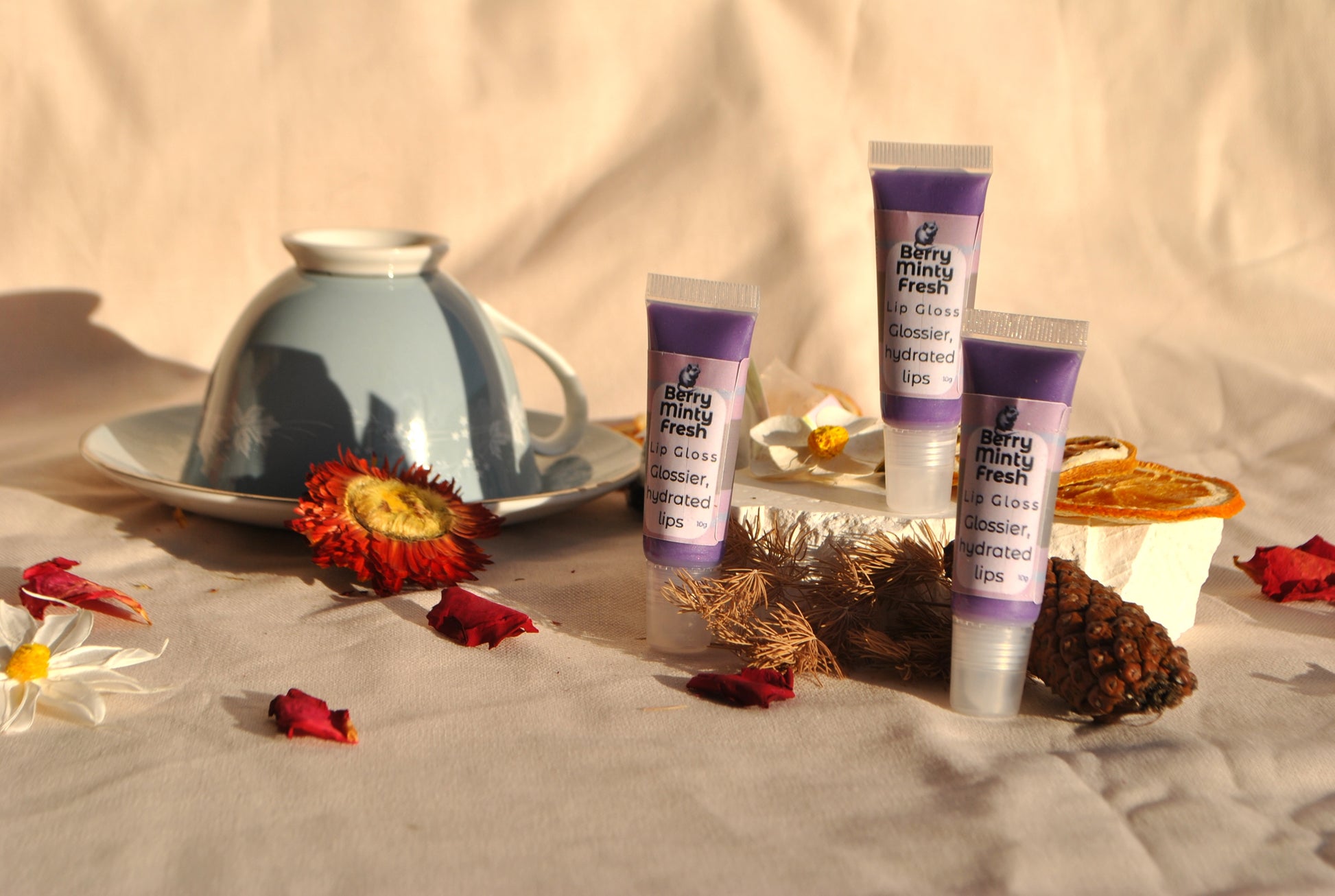 Three tubes of berry.minimum fresh lip gloss on a textured surface with a cup and flowers.