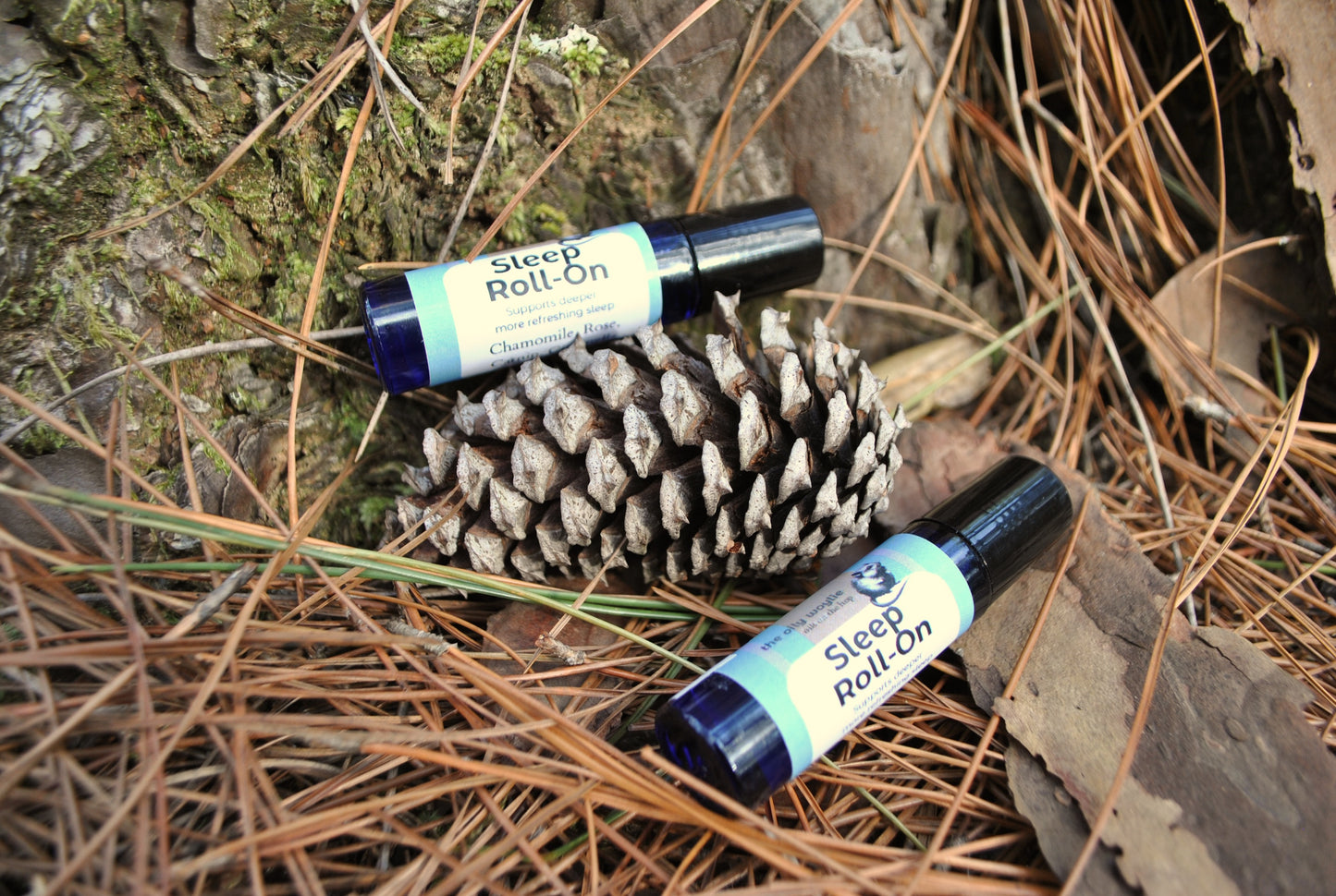 Two bottles of 'Sleep Roll On' on a pine cone and wooden surface with pine needles.