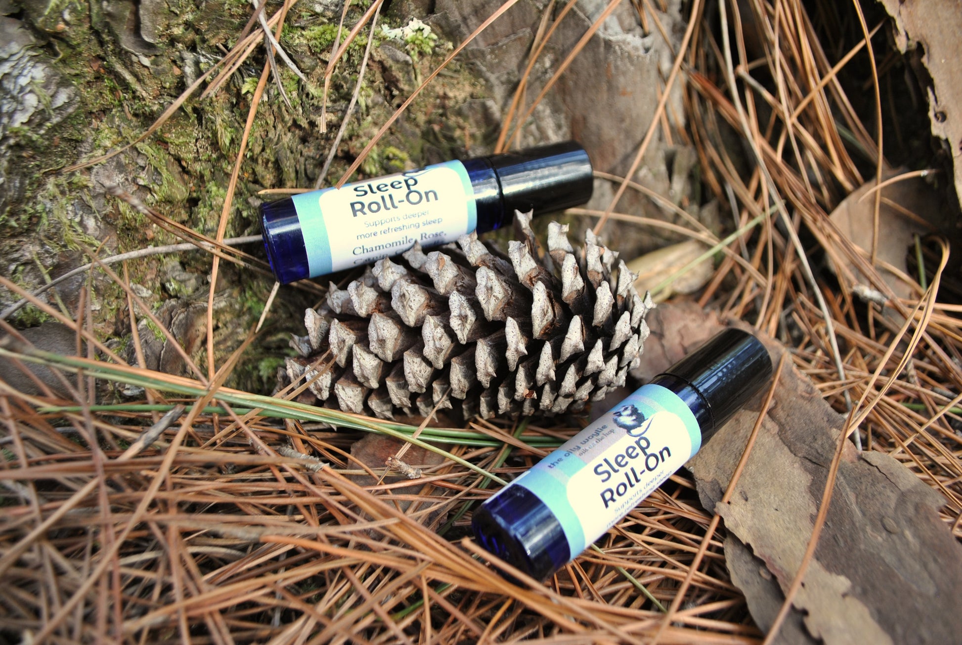 Two bottles of 'Sleep Roll On' on a pine cone and wooden surface with pine needles.