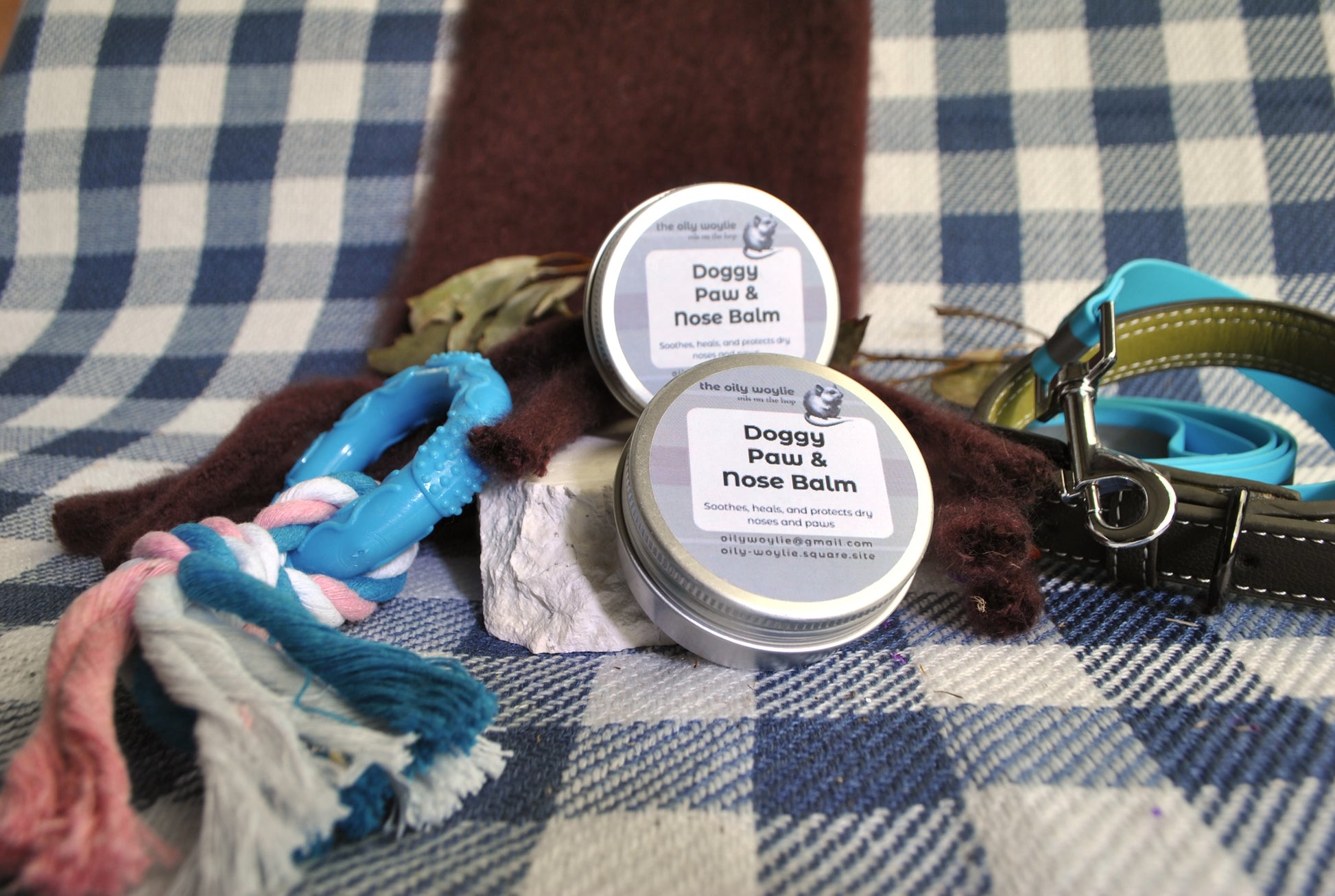 Two tins of Doggy Paw & Nose Balm on a checkered blanket with dog accessories.