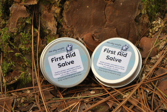Two tins of 'First Aid Salve' on a natural background with leaves and twigs.